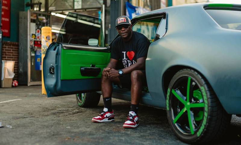 a man wearing an 'I love EPA' shirt sits in the front seat of a vintage blue car, facing outward, in a cap and sneakers