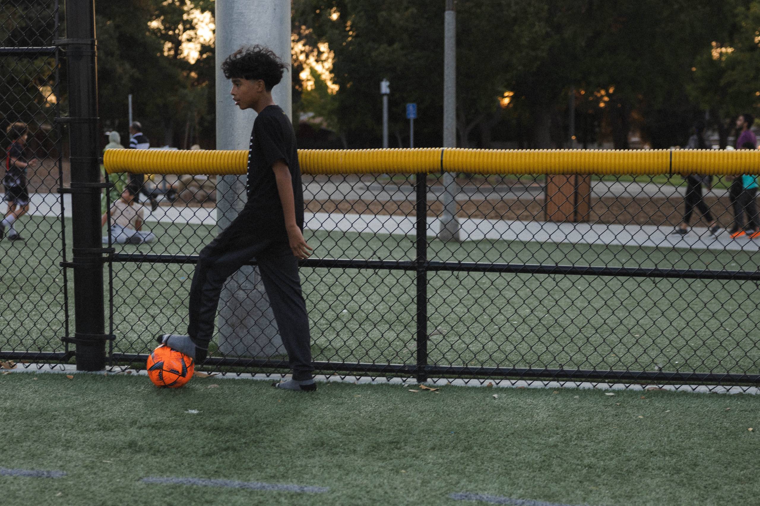 a teenage boy dribbles a soccer ball in his socks on the sidelines of a soccer field