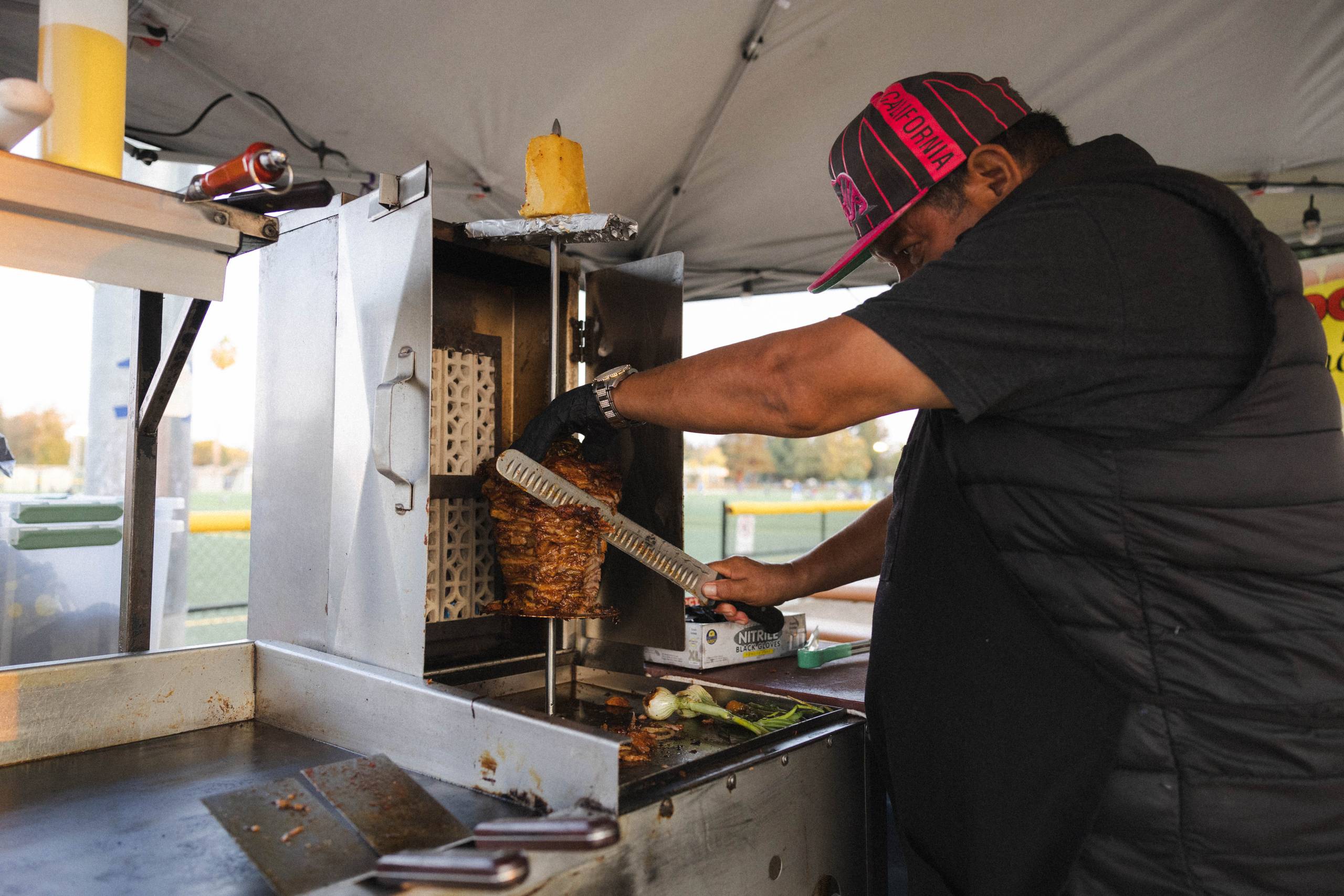 a taquero cuts al pastor meat from a spinning trompo during a local soccer game