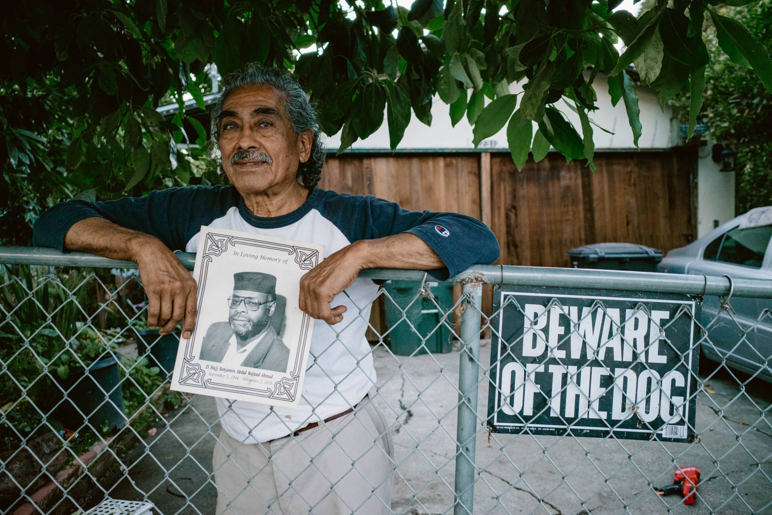 an elderly Latino man poses on the fence in front of his home in East Palo Alto