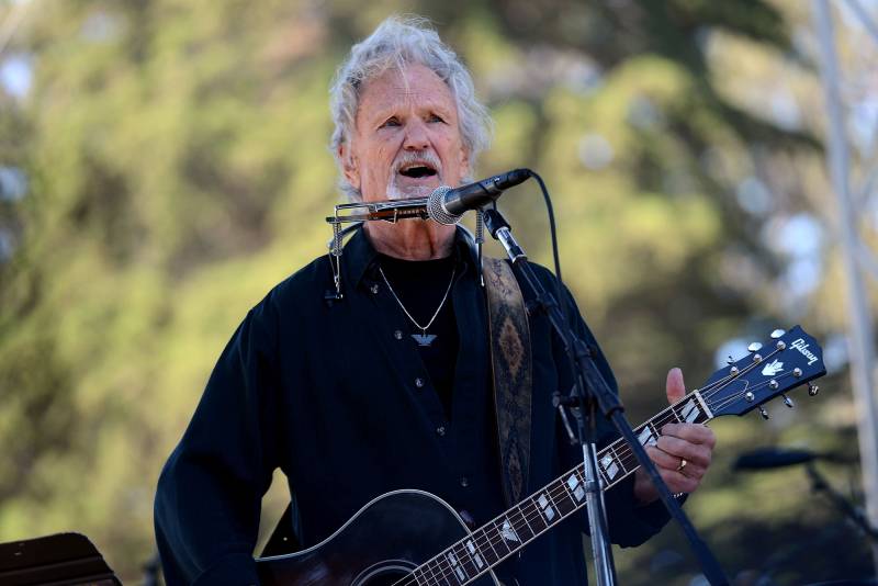 A man with greay hair in a black top plays the guitar and sings with trees as a backdrop