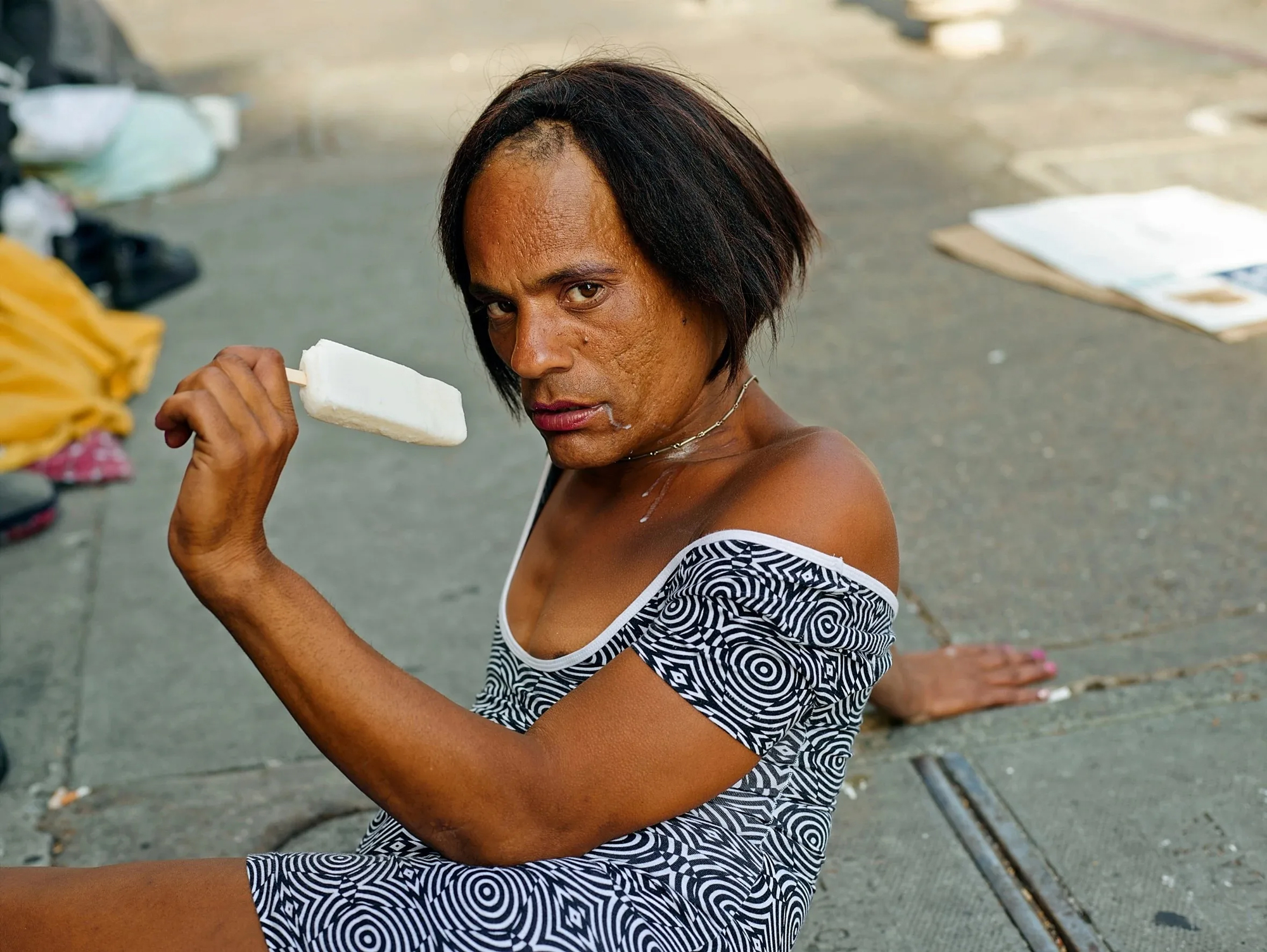 Person sits on sidewalk with popsicle in hand, looks directly at camera