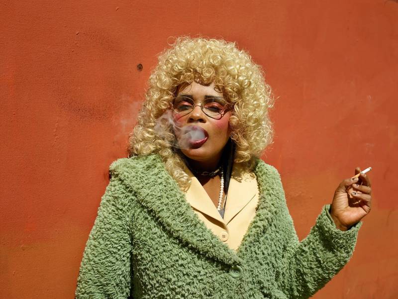 Person with curly blonde hair smokes cigarette in front of red wall