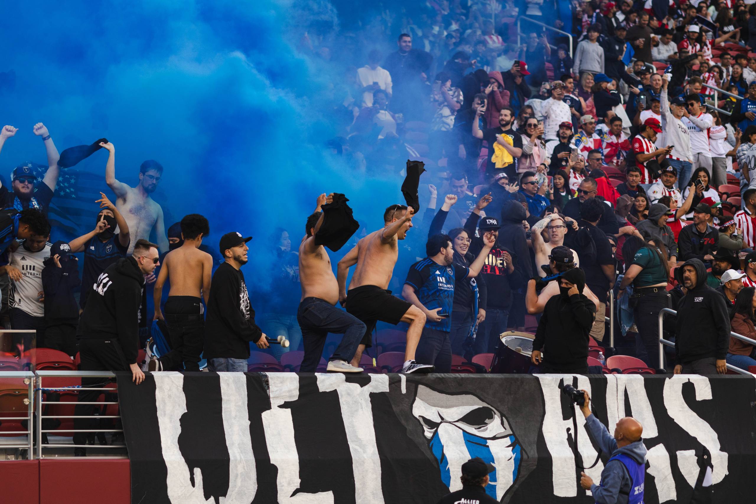 shirtless men cheer in stands with blue smoke behind them in stadium