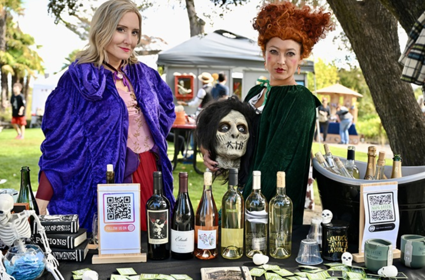 Two women in witchy costumes stand behind a table of wine bottles and Halloween decorations.