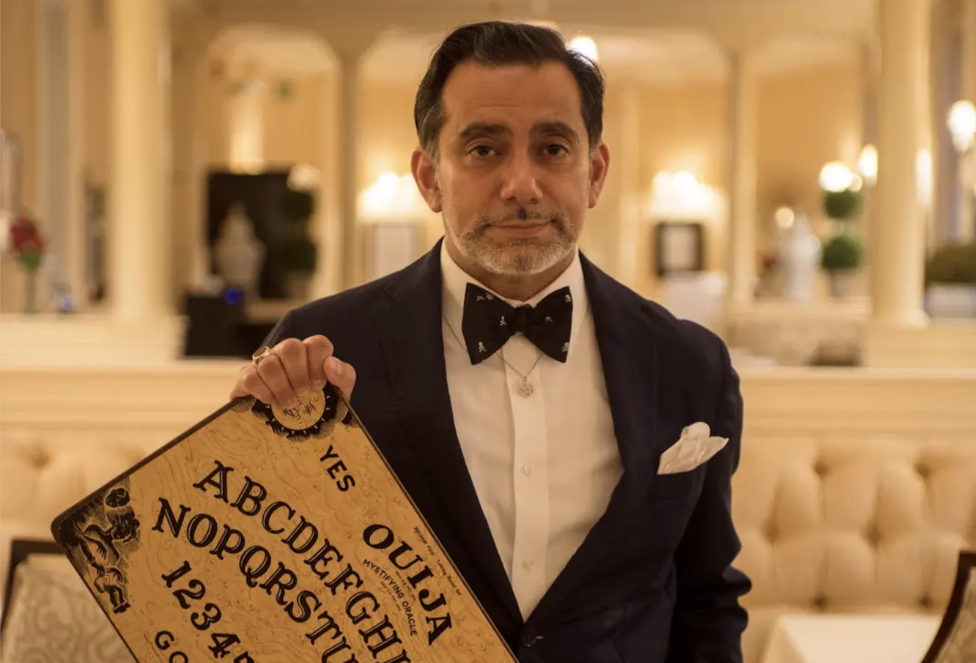 A man in a tuxedo stands in a grand dining room clutching a ouija board.