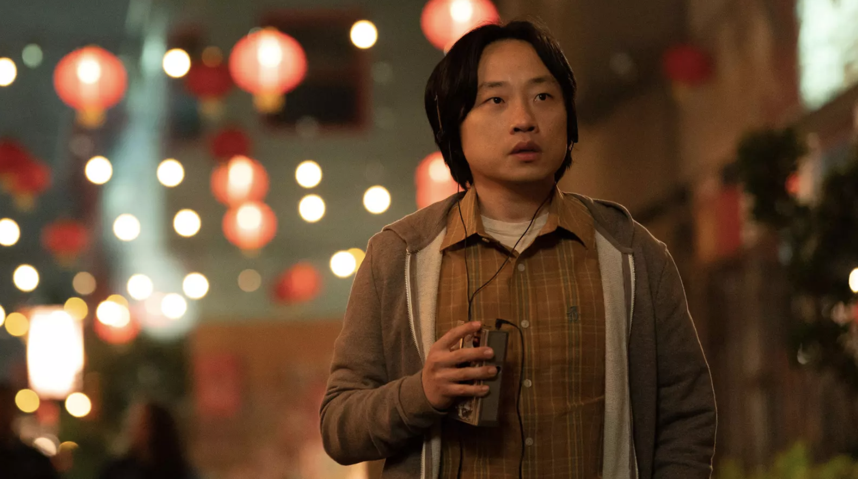 A young Asian man walks down a street at night, red lanterns and white lights hanging behind him. 