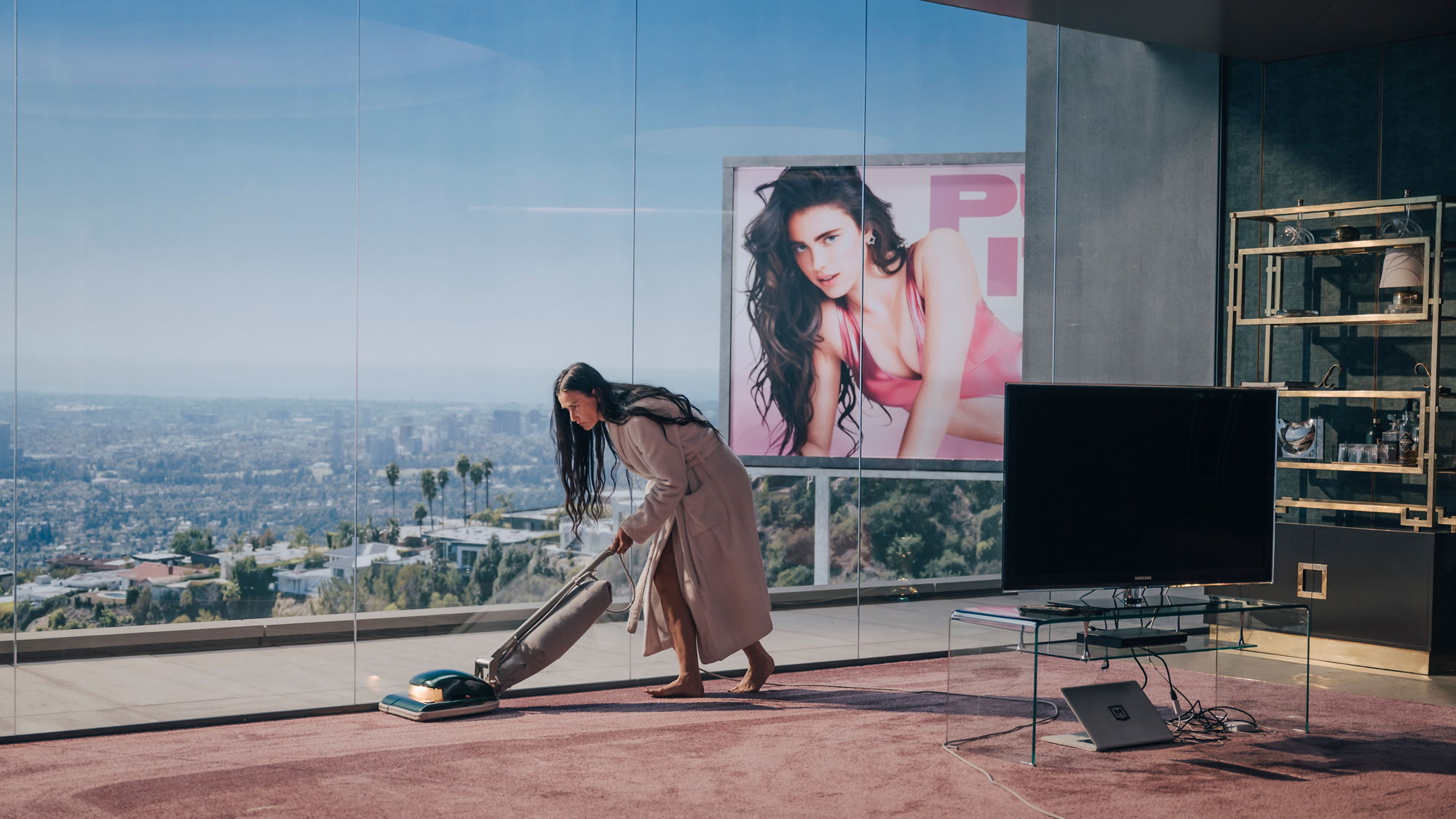 Woman vacuums in front of large windows, billboard of woman in leotard in background