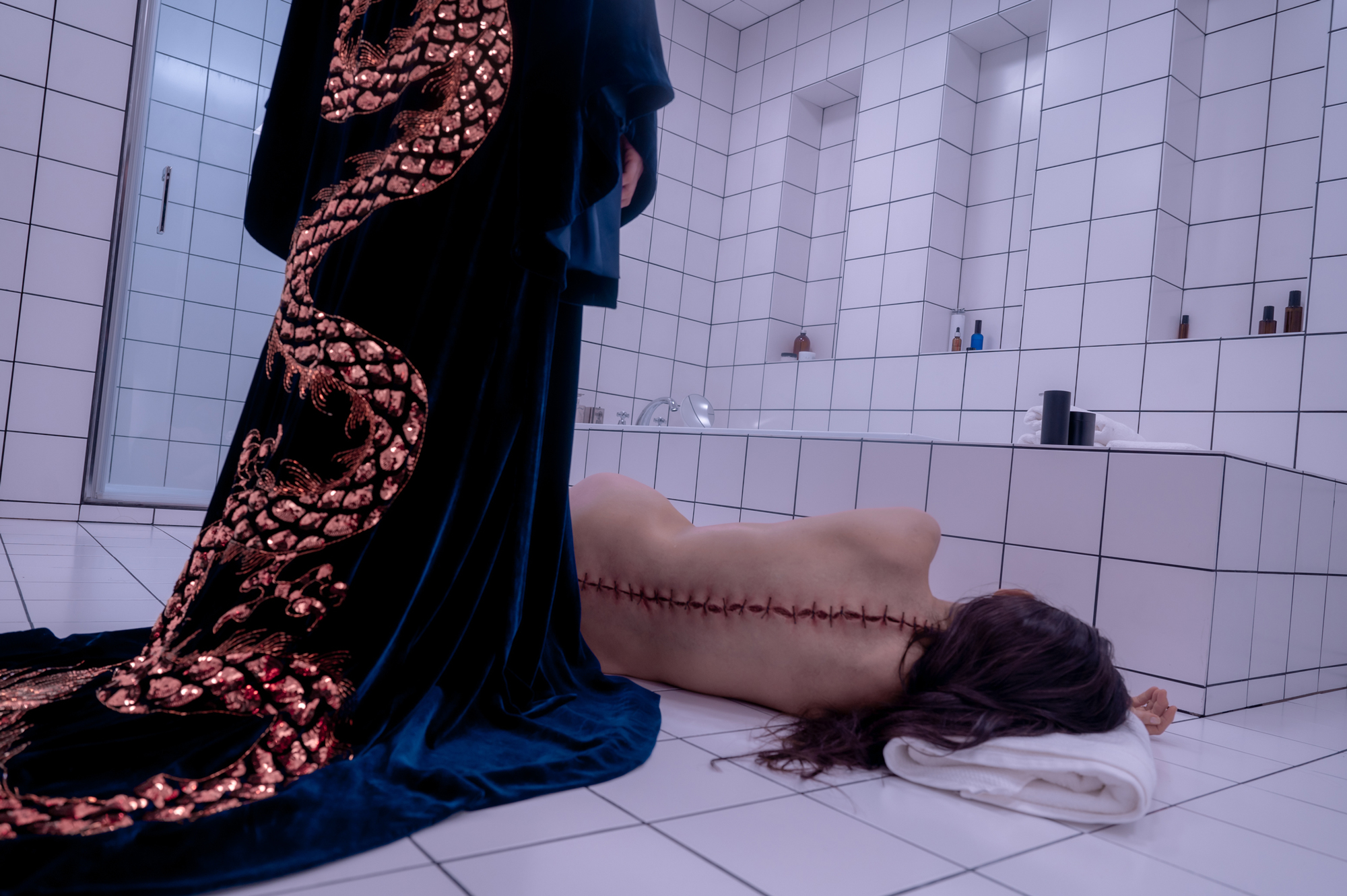 Person in robe with gold dragon pattern stands over white woman with sutures down spine in tiled room