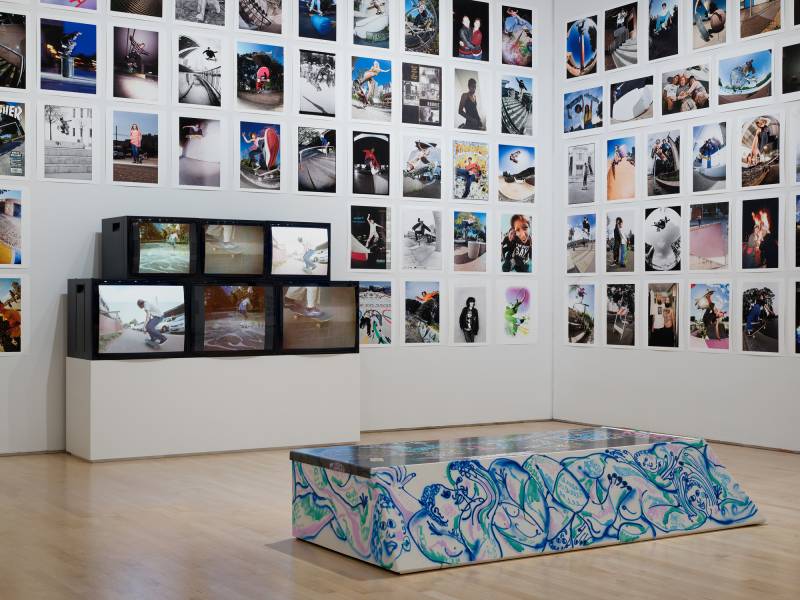 gallery with grid of photographs covering walls, six tvs and painted bench