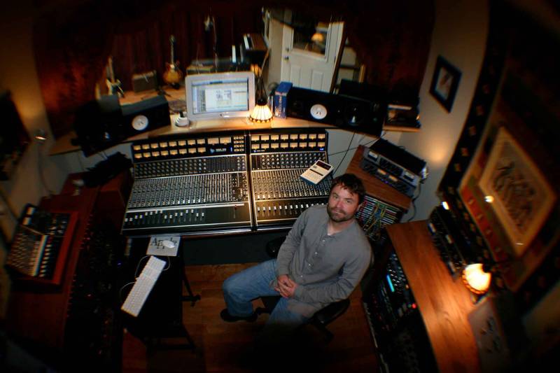 A man in a beard, button-down shirt and jeans sits at a mixing console, seen from above