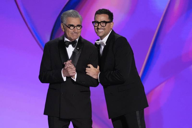 Two men, both wearing tuxedos and black rimmed spectacles, smile together on a stage.