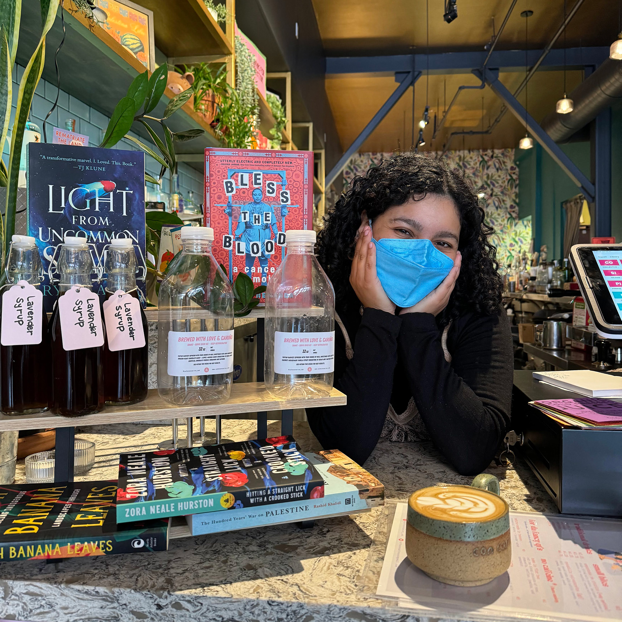 A coffee shop owner in a face mask poses for a portrait behind the counter.