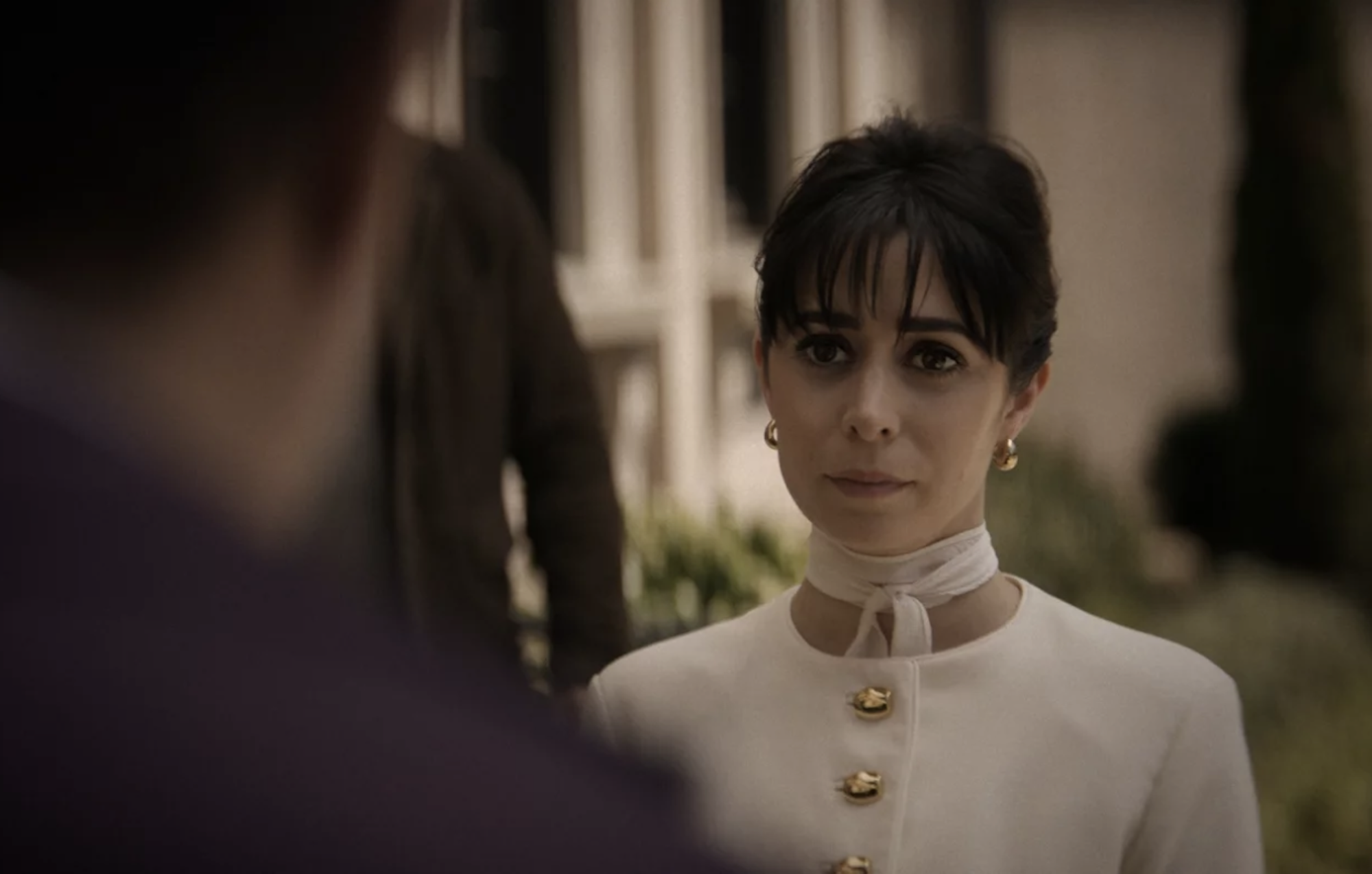 A woman with dark hair and pale skin stands in a courtyard wearing a smart white suit