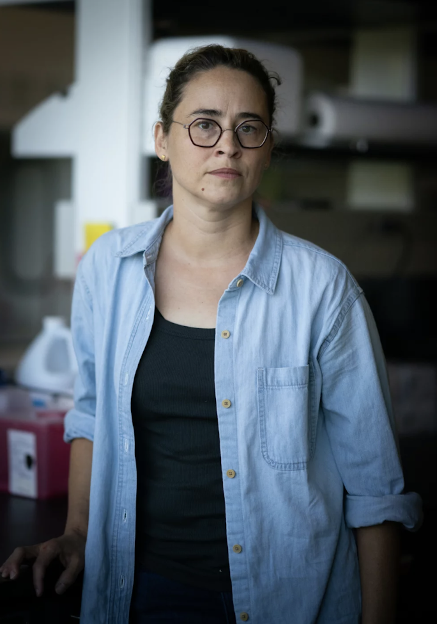 A white woman wearing round glasses stands in a room, looking serious.