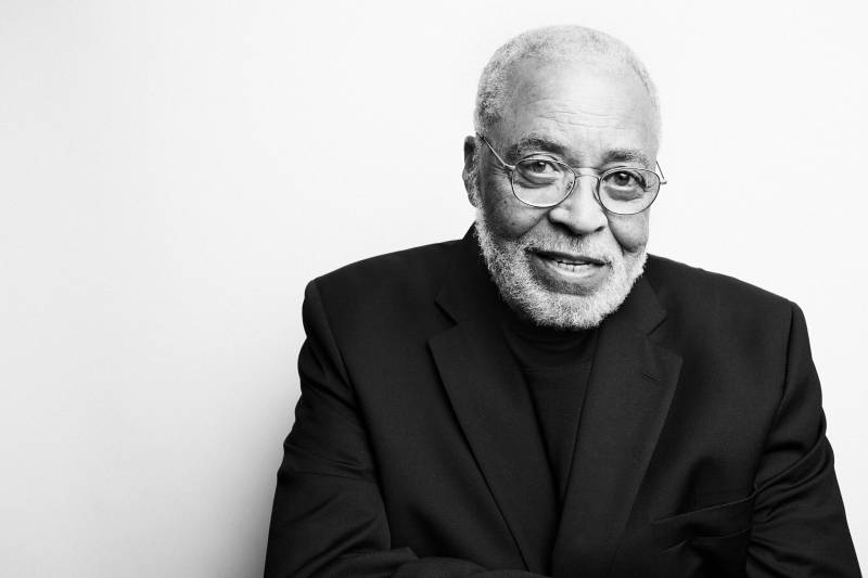 A portrait of a senior Black man with a white beard. He is wearing a black suit and round glasses.