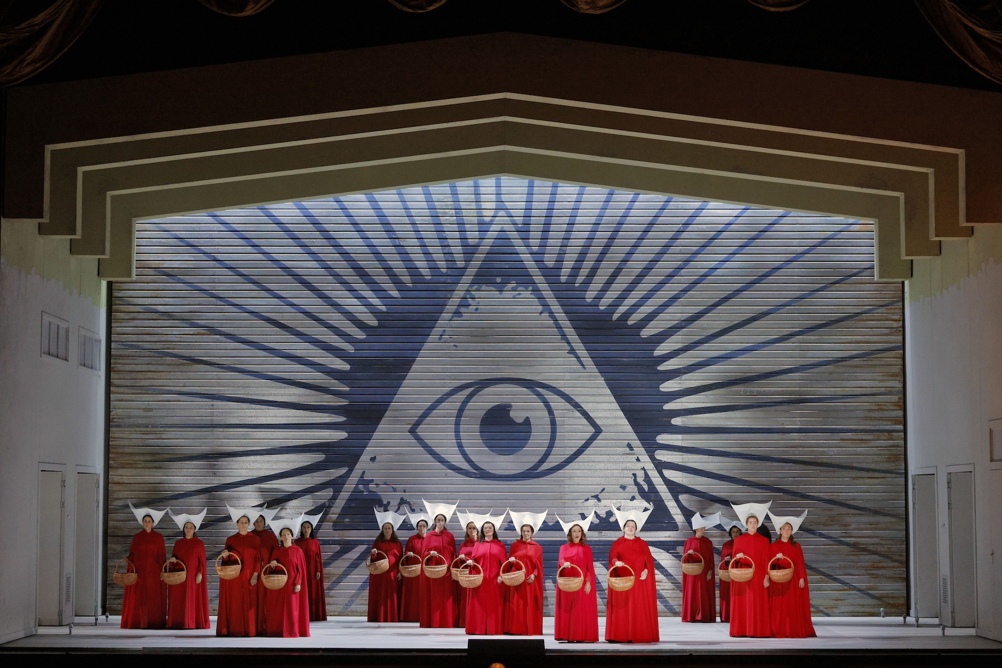 A stage set featuring a backdrop decorated with a large triangle and eye. On stage are 18 women all dressed in long red cloaks.