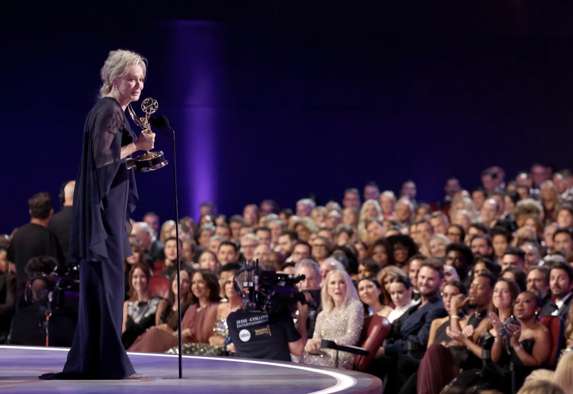 A mature woman in an evening gown stands on stage holding an award before a large audience.