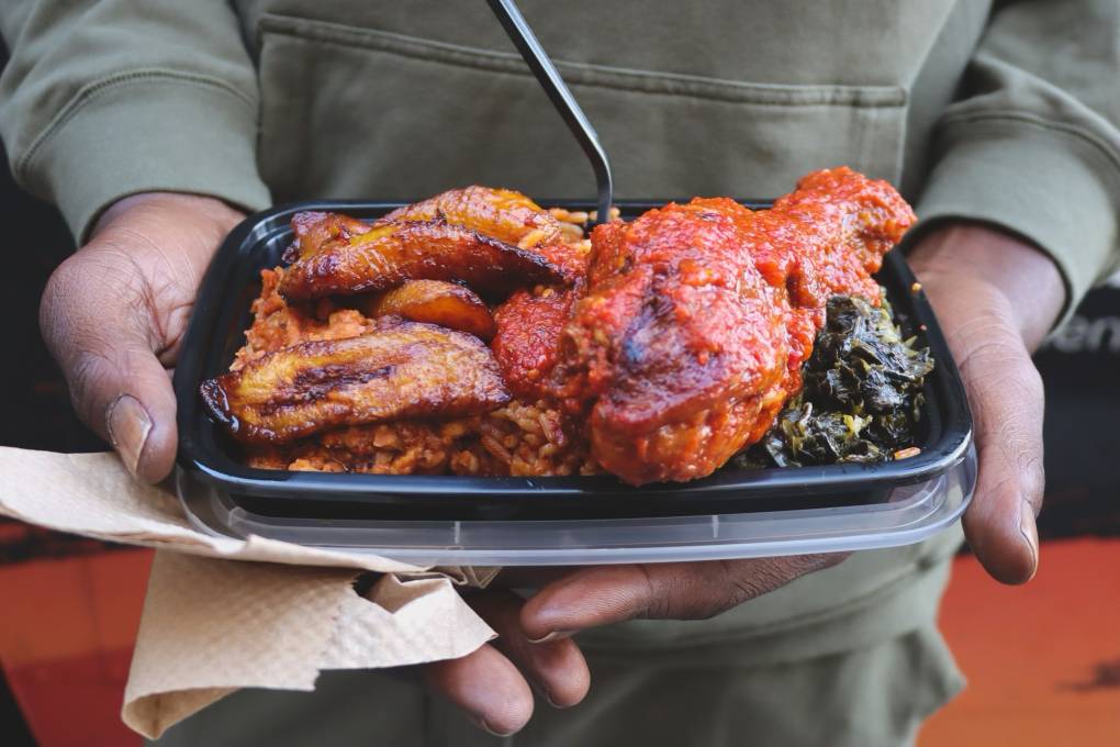 Man holding a takeout container of birght orange jollof rice with chicken and plantains.