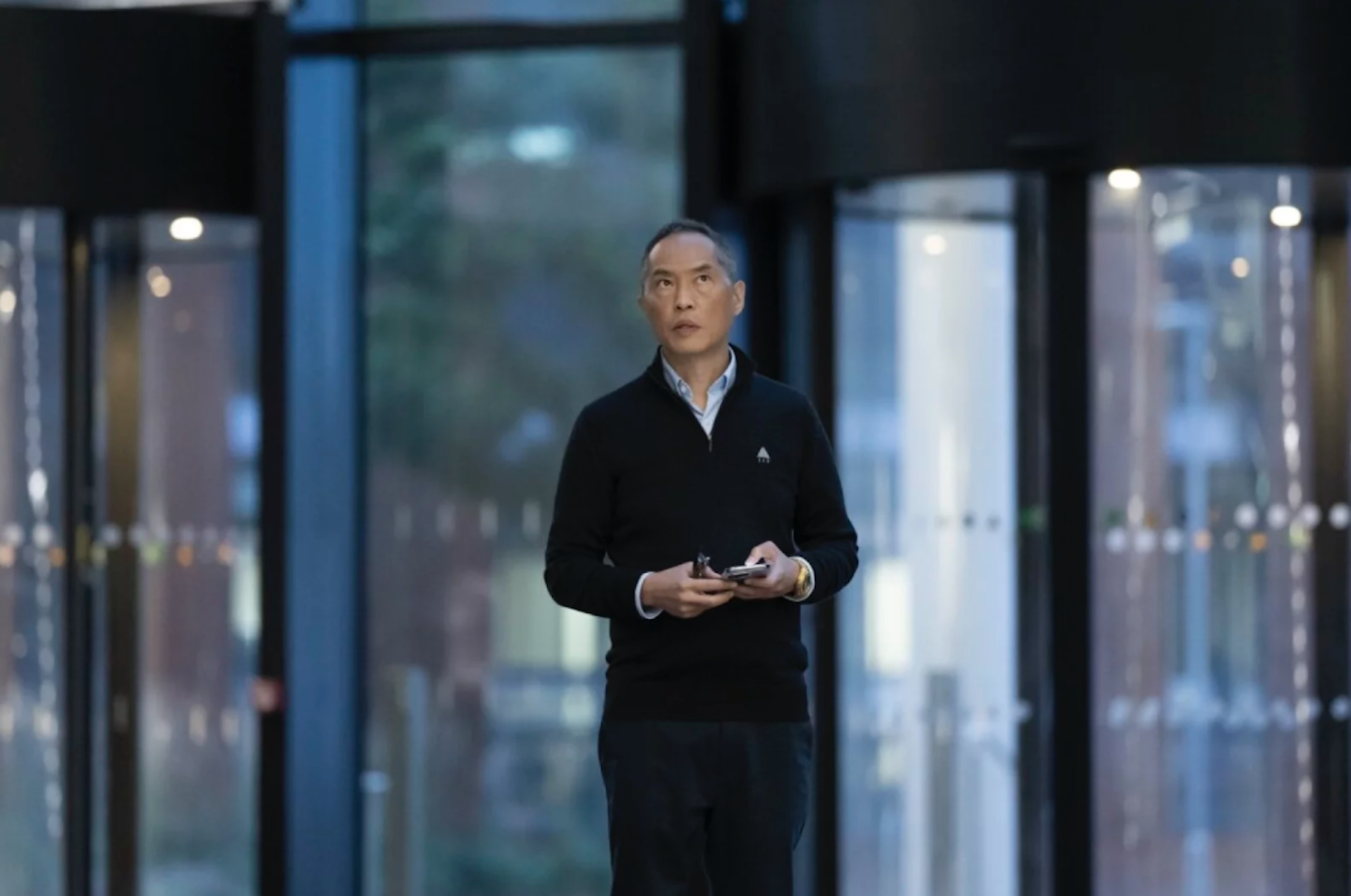 A middle aged Asian man stands in the lobby of a glass office building.