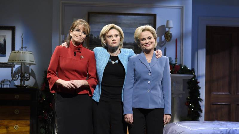 Three white women wearing pant suits stand side by side in living room set.