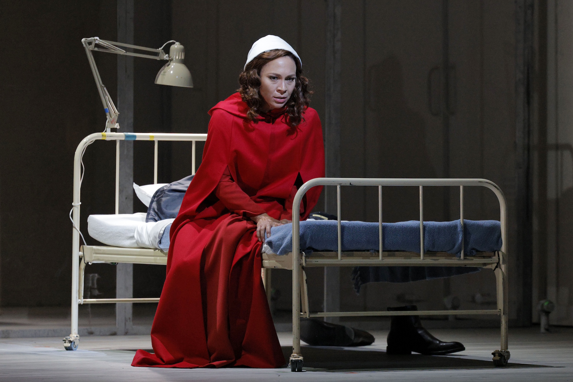 A perturbed looking woman wearing a red cloak and white bonnet sits on a twin bed.