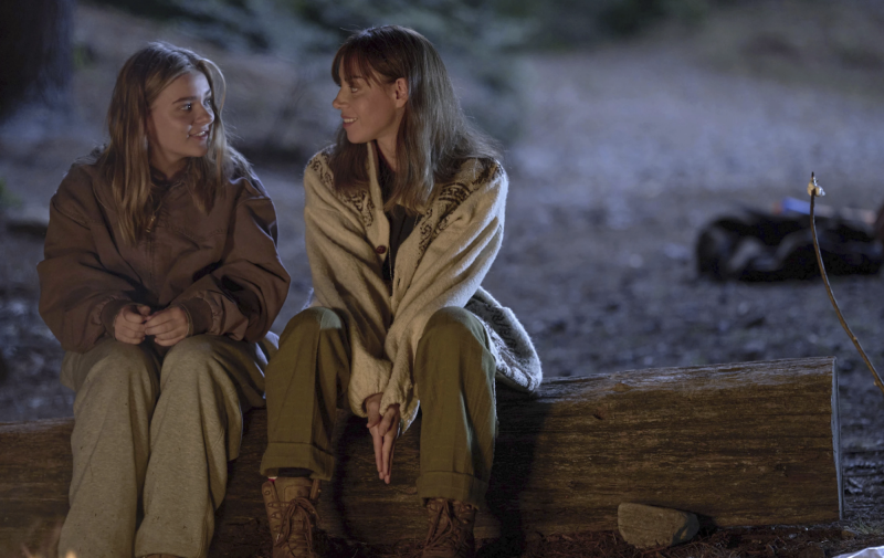 Two attractive white women sit on a log, outdoors at night, looking fondly at each other.