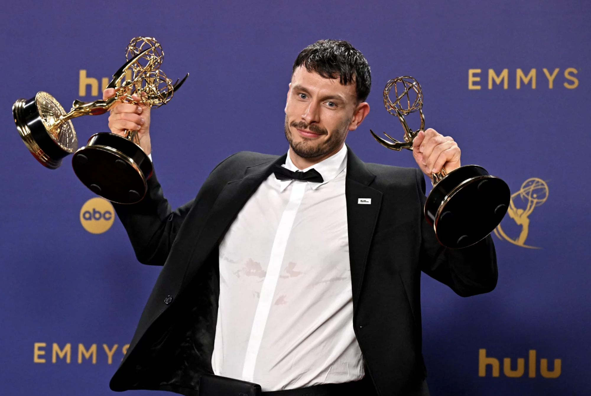 A white man in a tuxedo holds up two awards triumphantly.