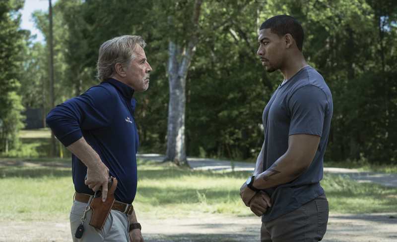 A older white man with one hand on his gun holster faces off with a younger Black man whose hands are folded in front of him. They are standing in a rural area.