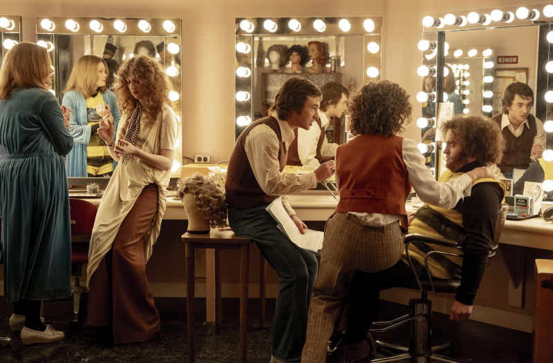 A group of men and women talk amongst themselves before mirrors in a dressing room.