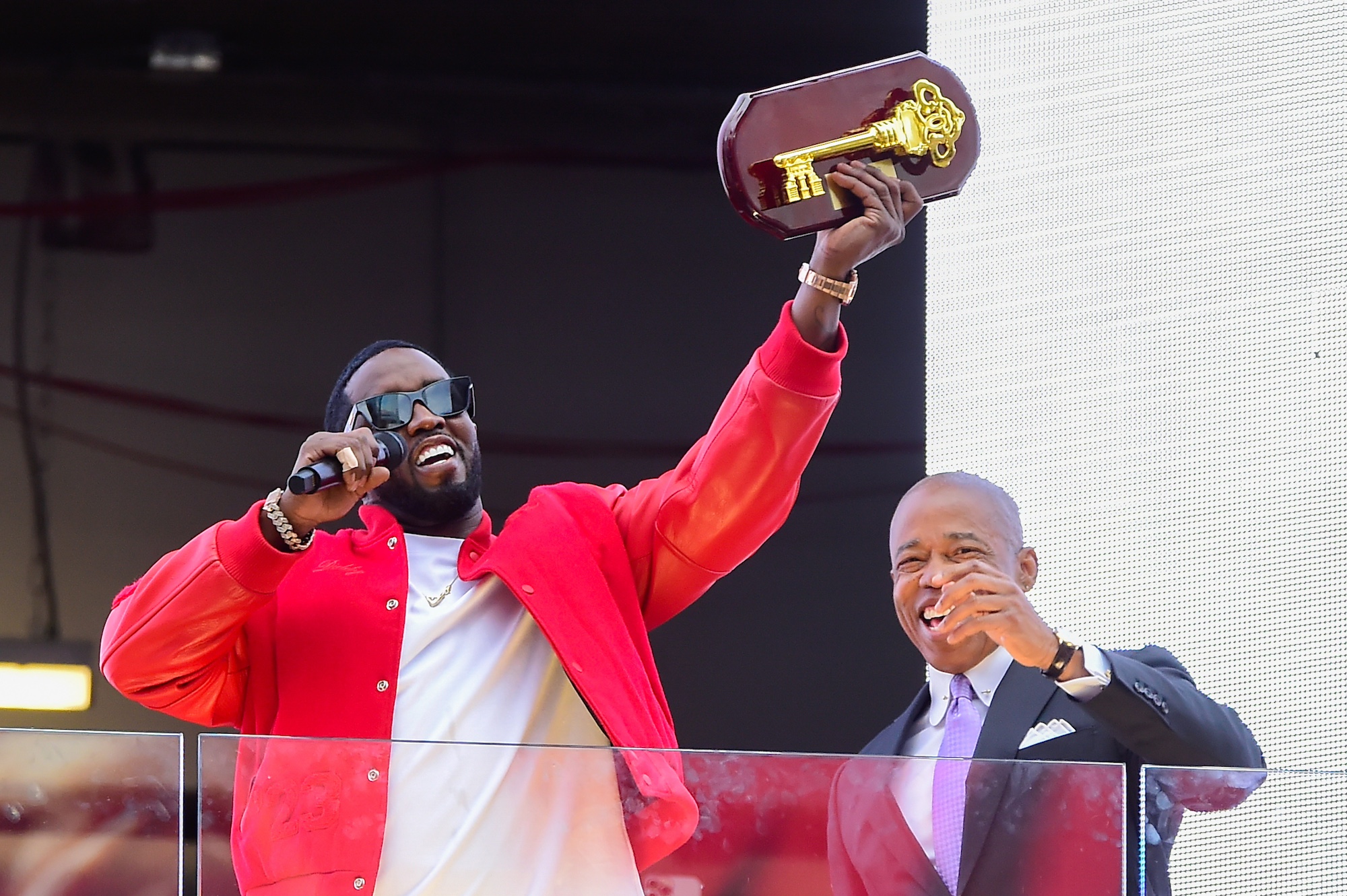 A Black man stands on a stage holding a microphone in one hand and a gold key mounted to a plaque in the other.