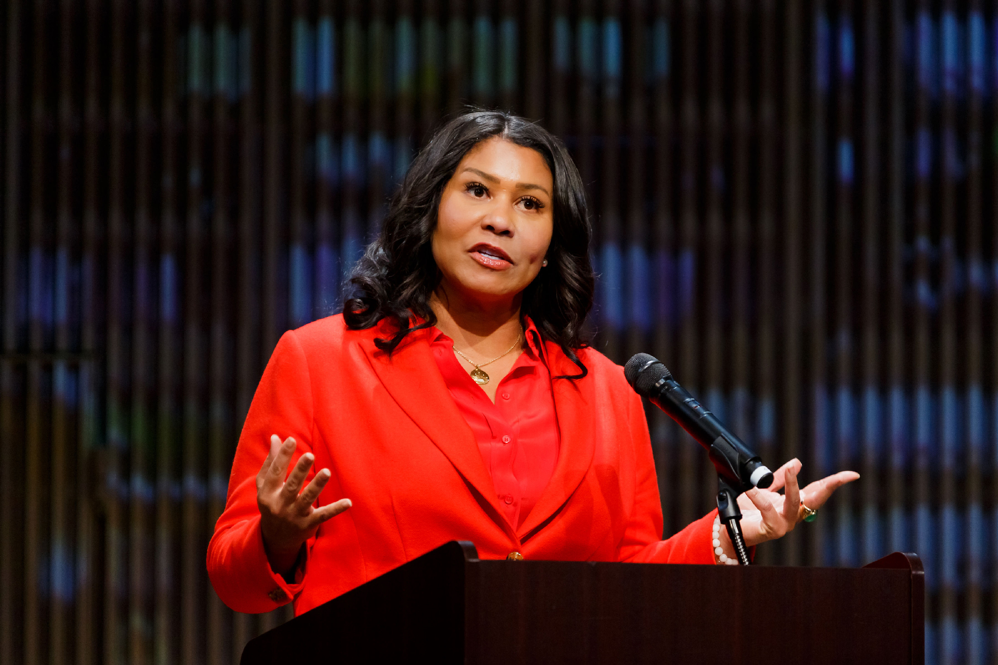 Black woman in red suit gestures with both hands at podium