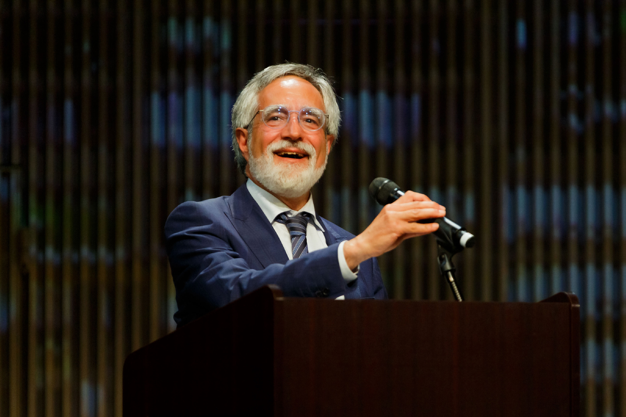 older white man with beard holds mic with one hand, smiling