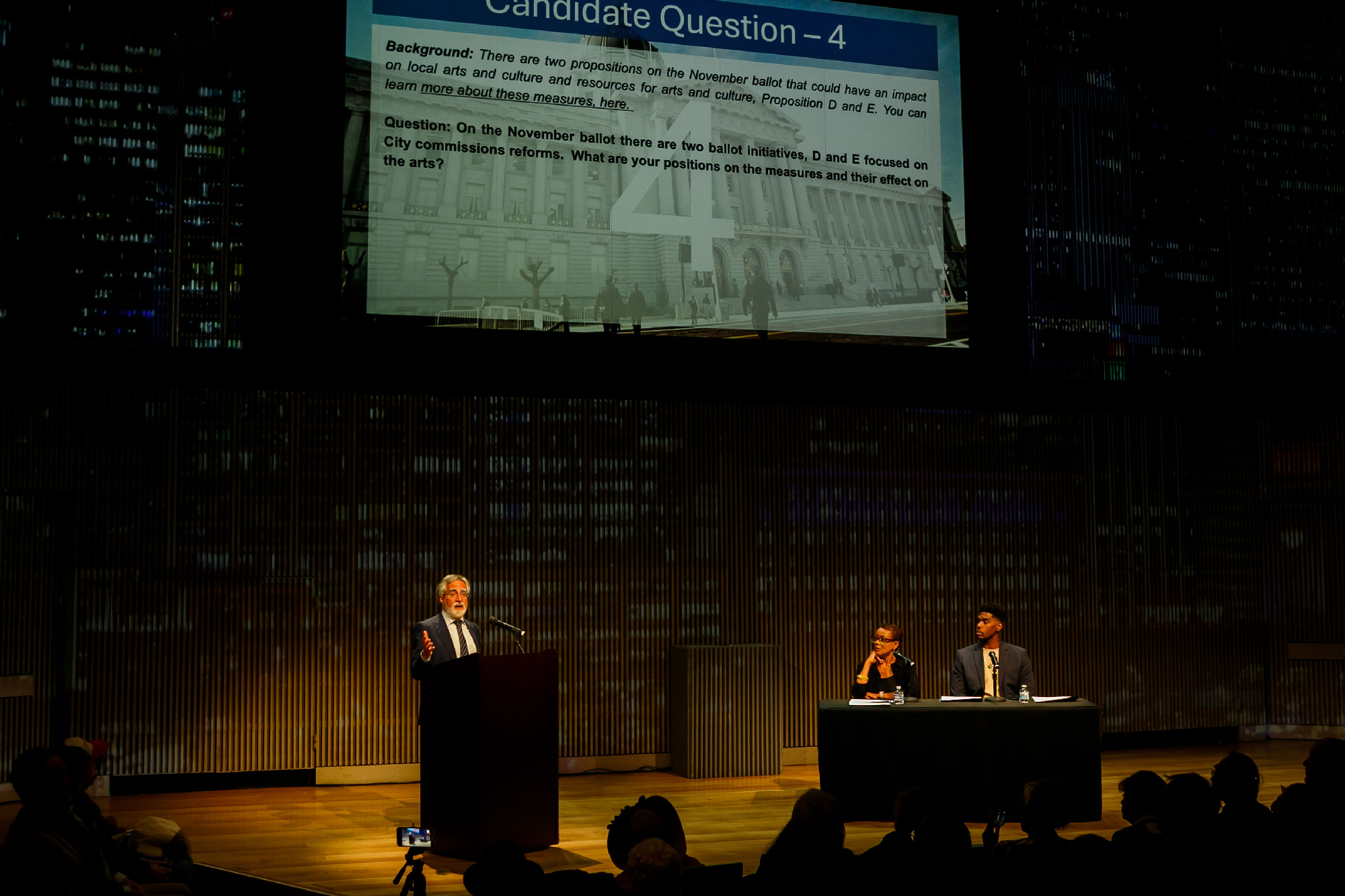 wide view of darkened stage with man at podium, two moderators at separate table and projected question above