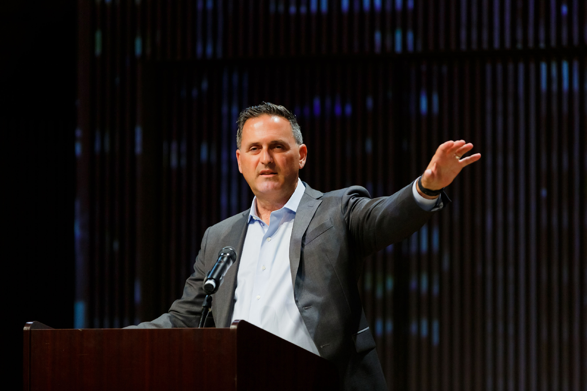 Man in suit without tie gestures with arm at podium