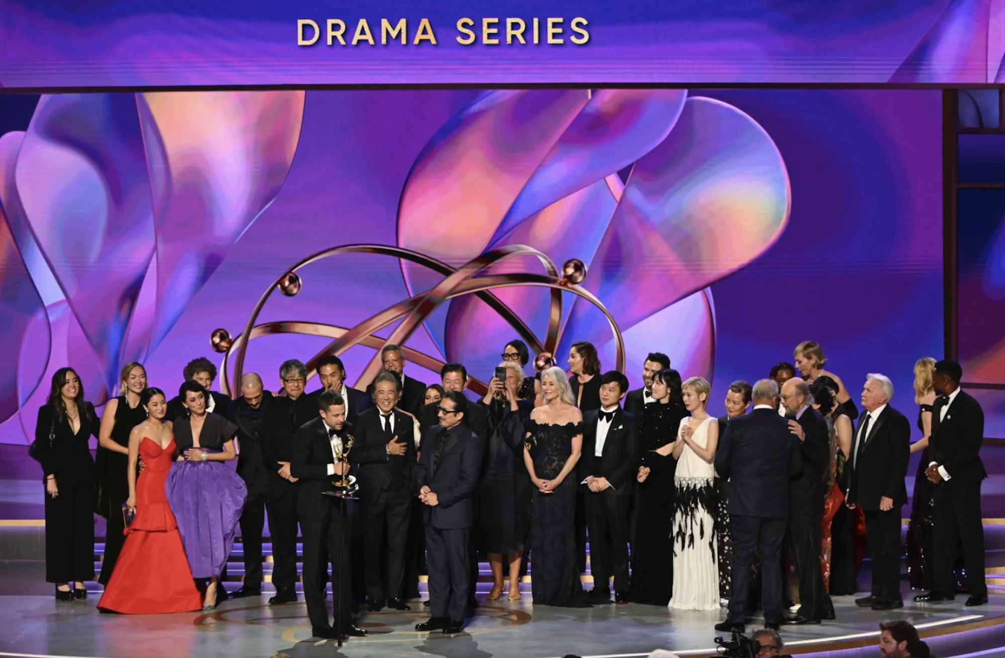 A large group of men and women in evening wear on an awards stage.