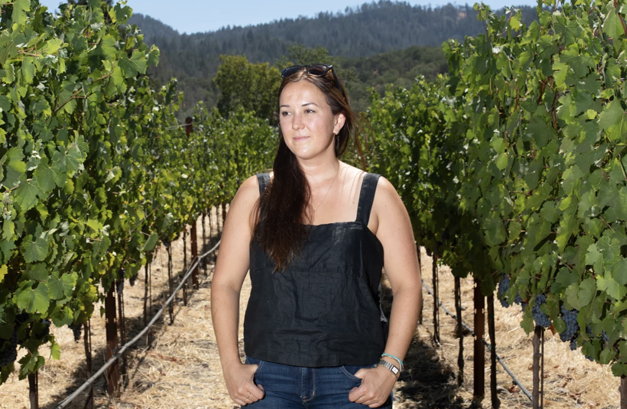 A white woman with long brown hair stands in the middle of a vineyard.