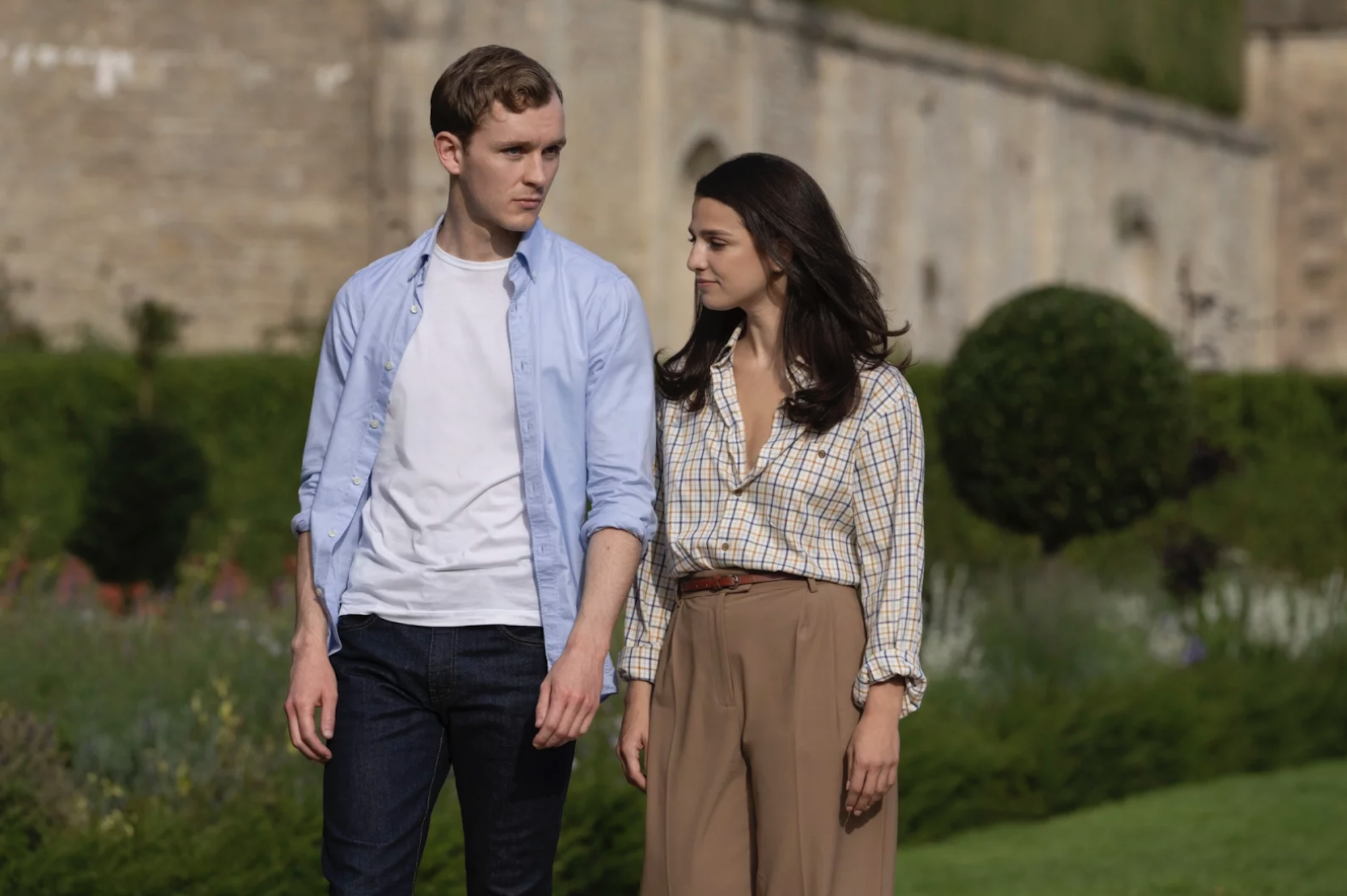 A white man and woman walk the grounds of a stately home.