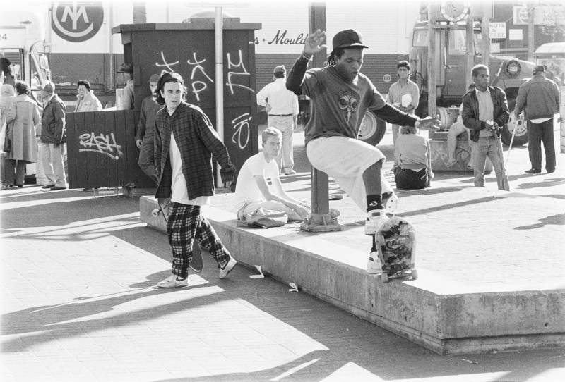 A skateboarder grinds on a concrete platform, while another watches on. Foot traffic is all around them.