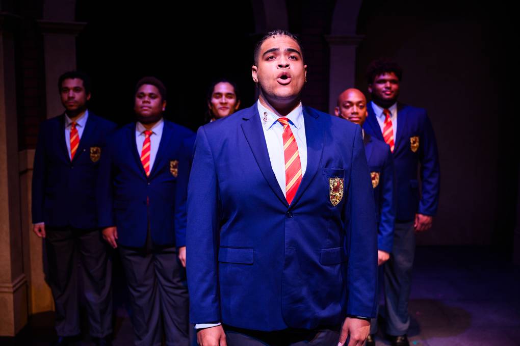 A young Black male student in a blue private school uniform with a red striped tie sings in front of a backing choir of four, similarly dressed.