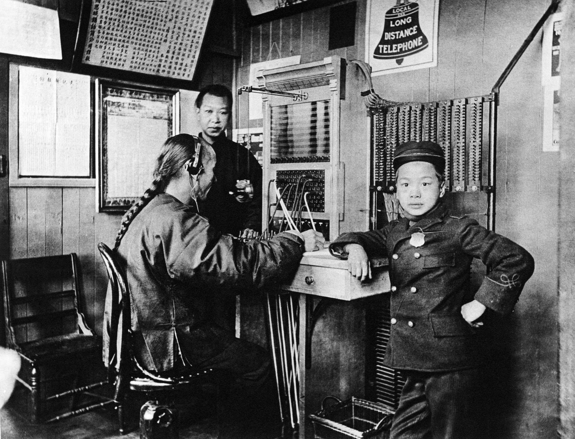 A man with a long braid dressed in traditional Chinese clothing sits at a switchboard with headphones on. A middle-aged man is standing near him, as is a small boy in an outfit that resembles a bell hop. 