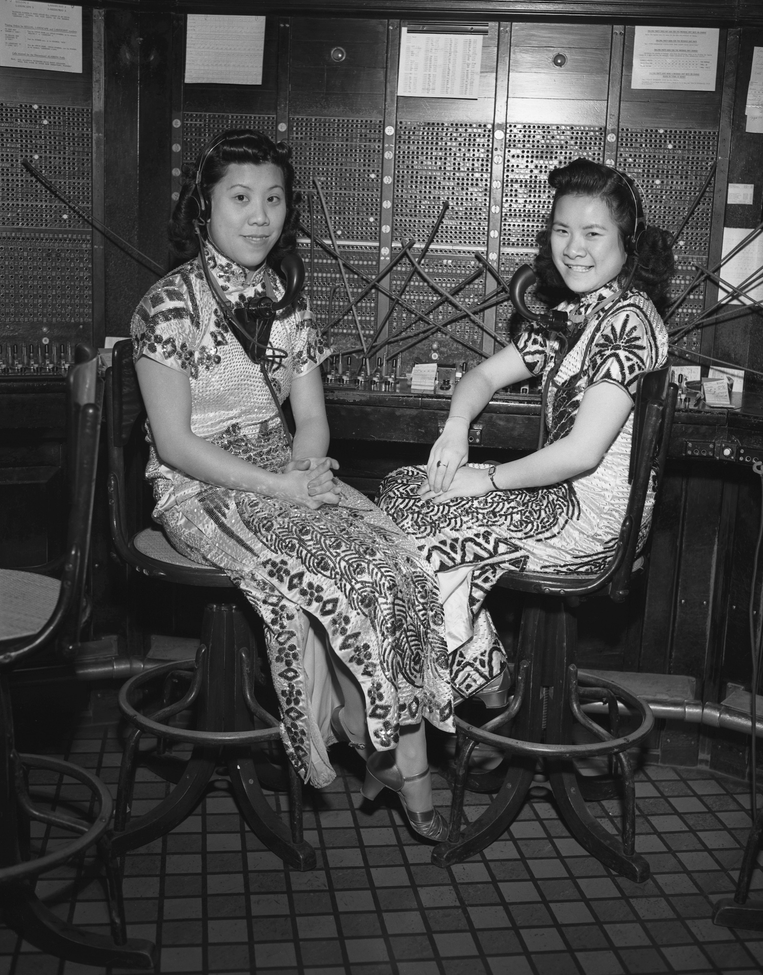 Two Asian women wearing intricate gowns sit in front of a switchboard.