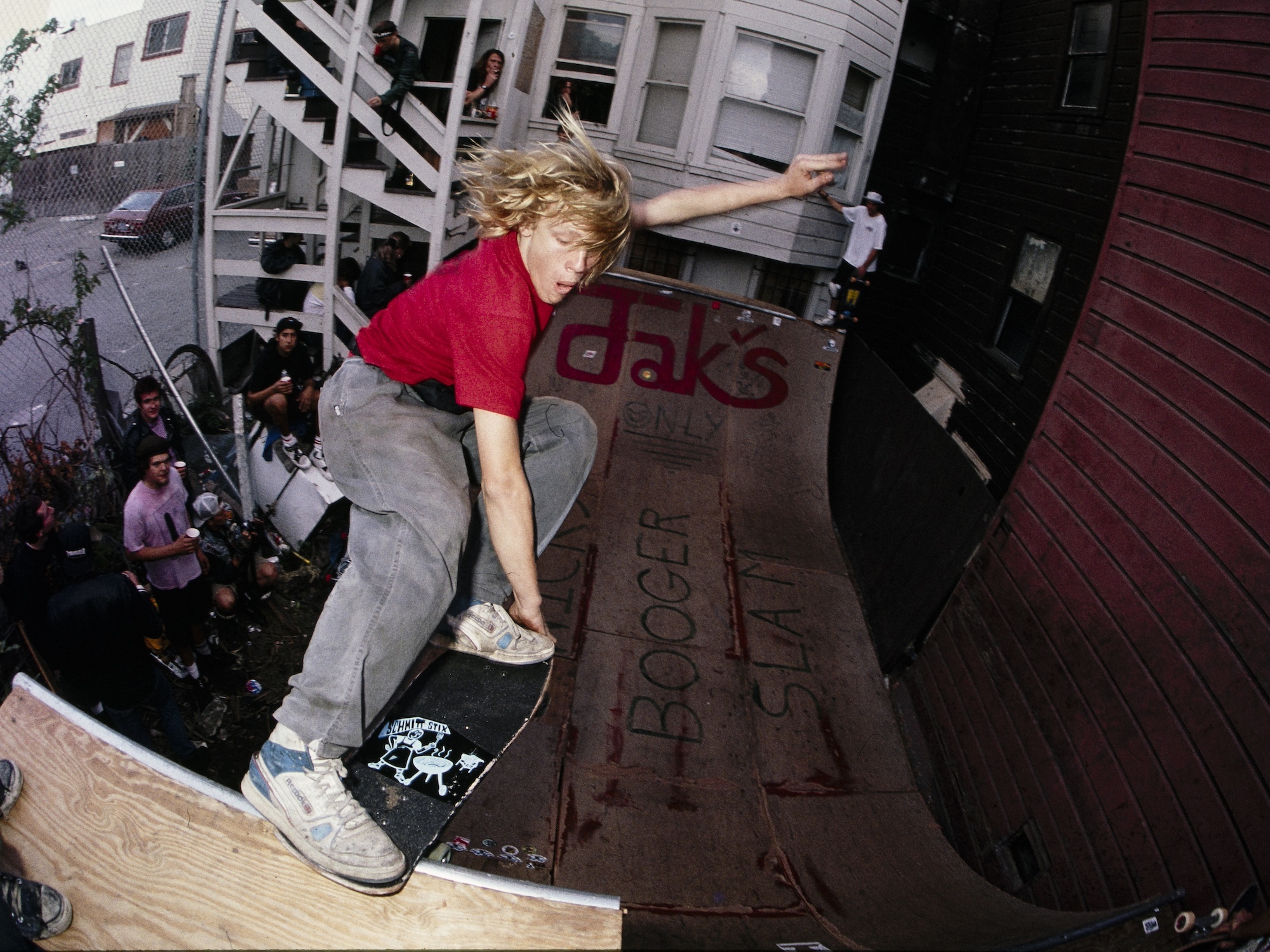 A young man balances on his skateboard on the top edge of a half pipe built behind someone's house. Spectators watch on from underneath.