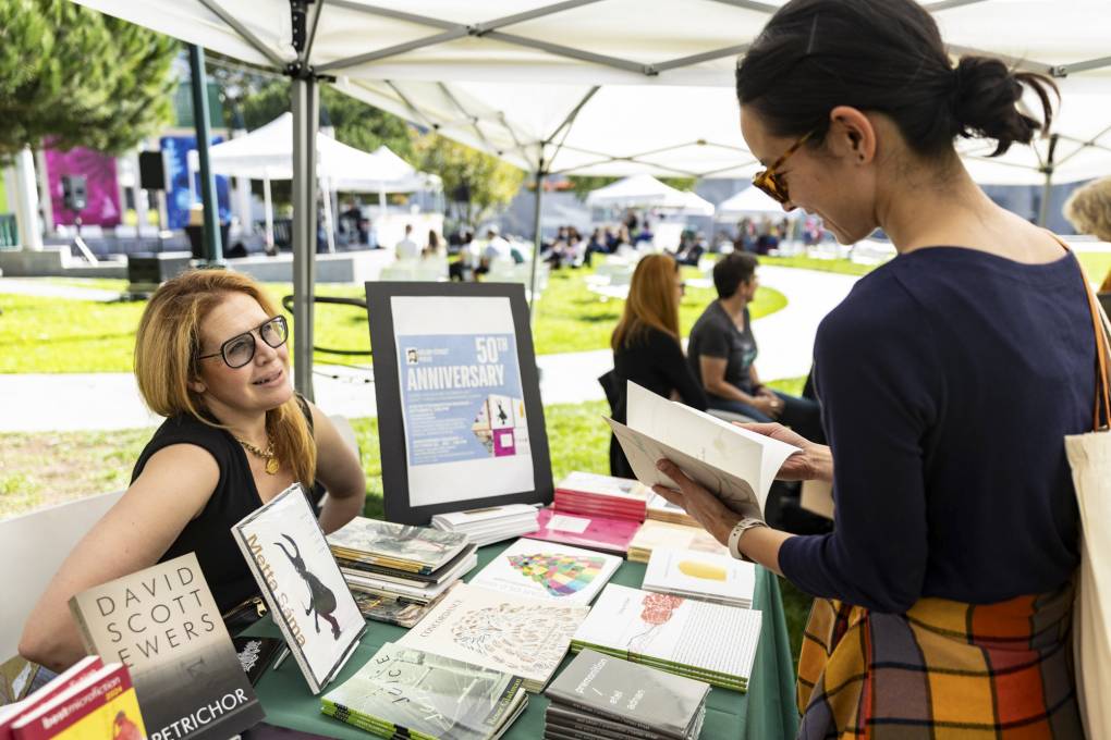 one person sits behind table of books while standing customer holds a book