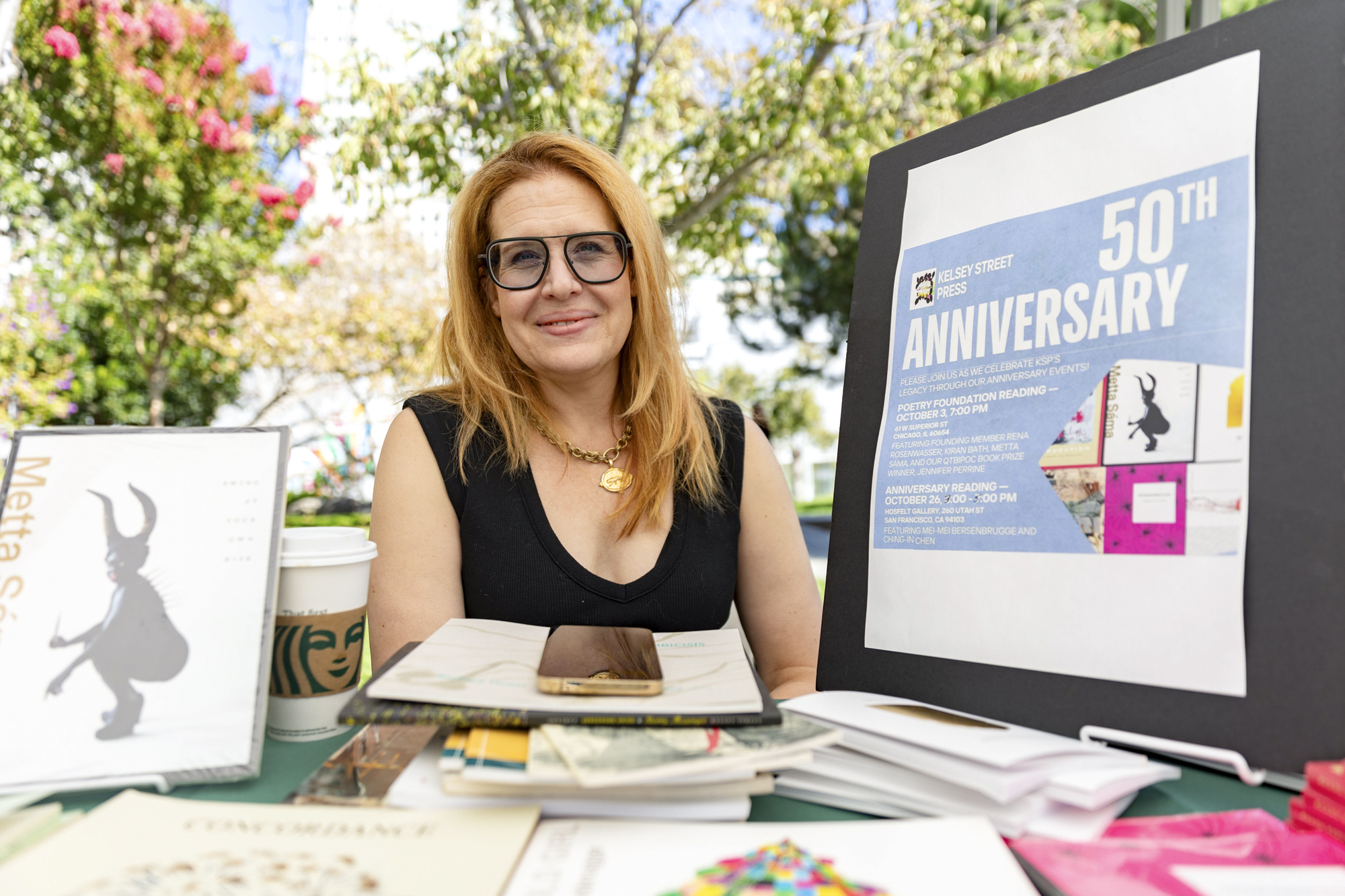 person sits behind table of books, smiling