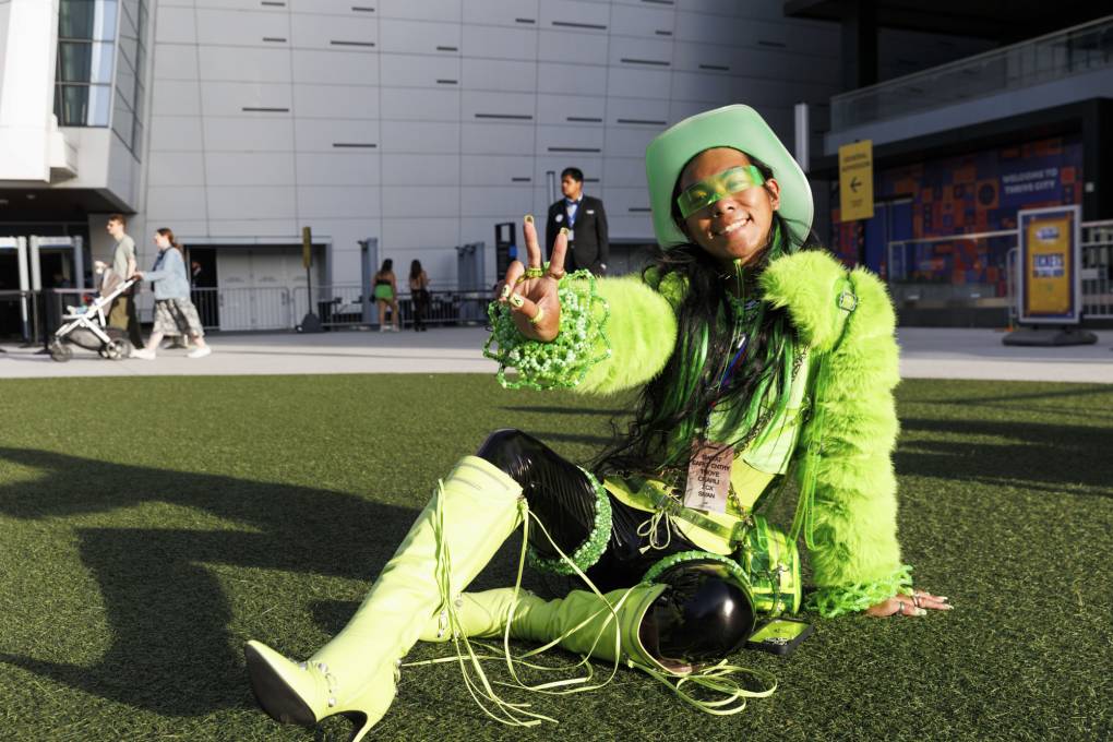 A woman wearing neon green boots, sunglasses, a hat and a jacket gives the peace sign with her hand while sitting outside on the ground.