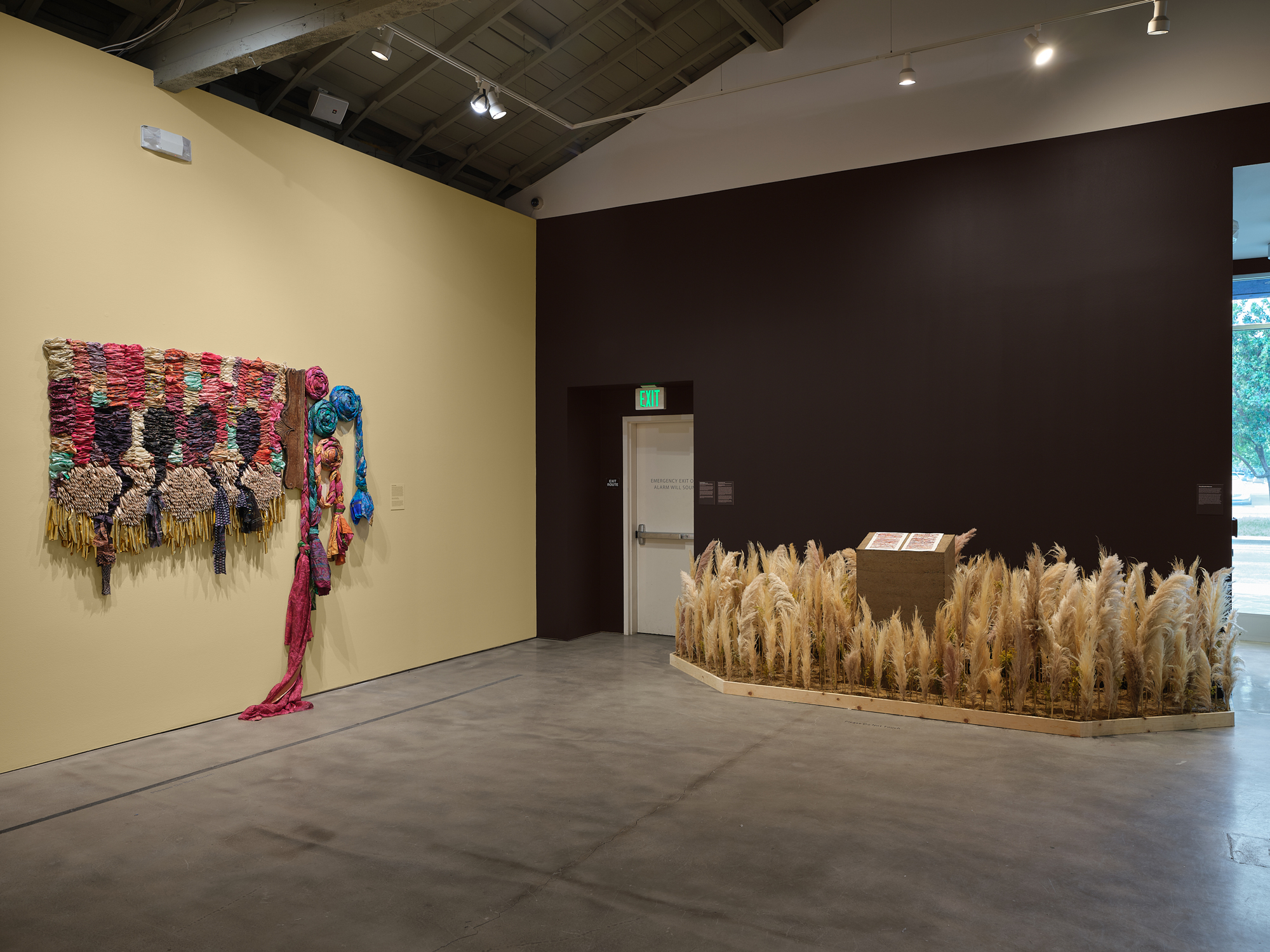 gallery view with textile piece on light yellow wall and pedestal in installation of dried grasses at right