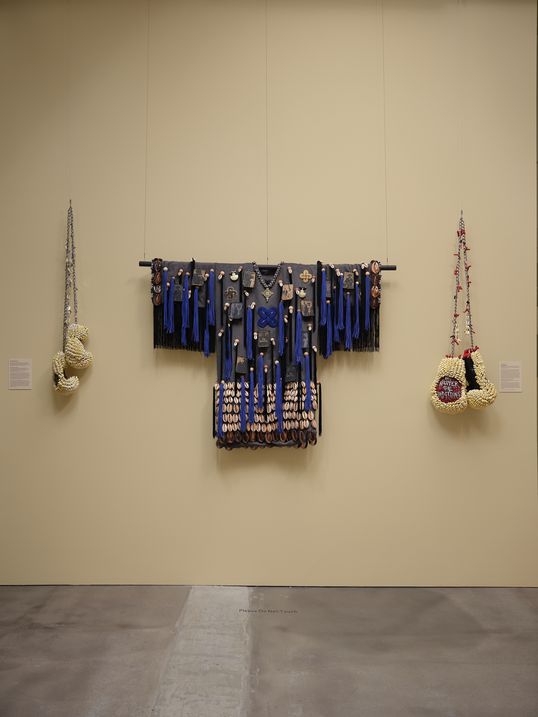 three sculptures on light yellow wall, two are embellished boxing gloves, one an embellished robe