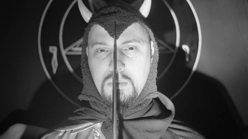 A man in a horned headdress, moustache and goatee poses before a pentagram, holding a sword that bifurcates his unsmiling face.