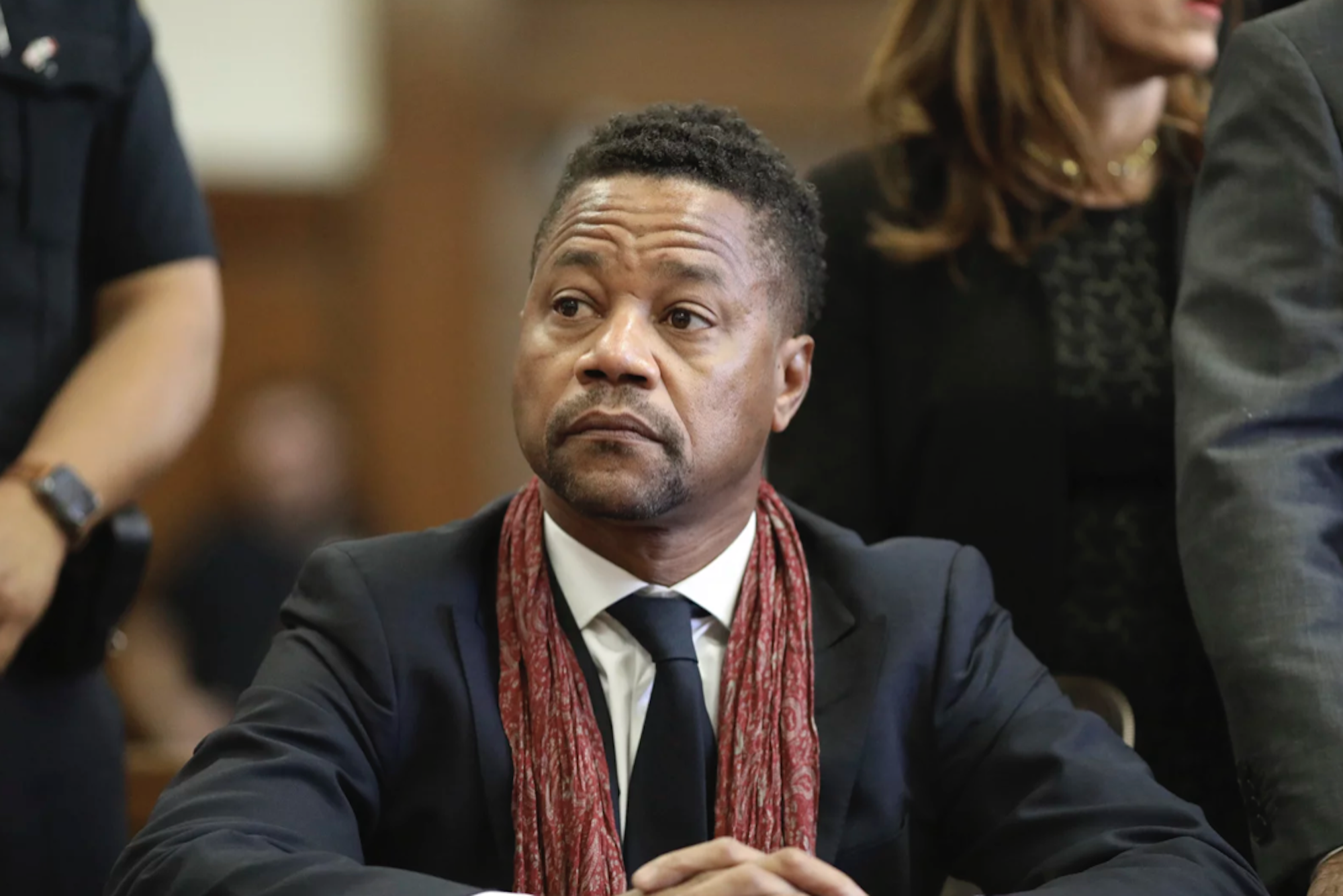 A Black man sits in court wearing a suit and red scarf, looking concerned.